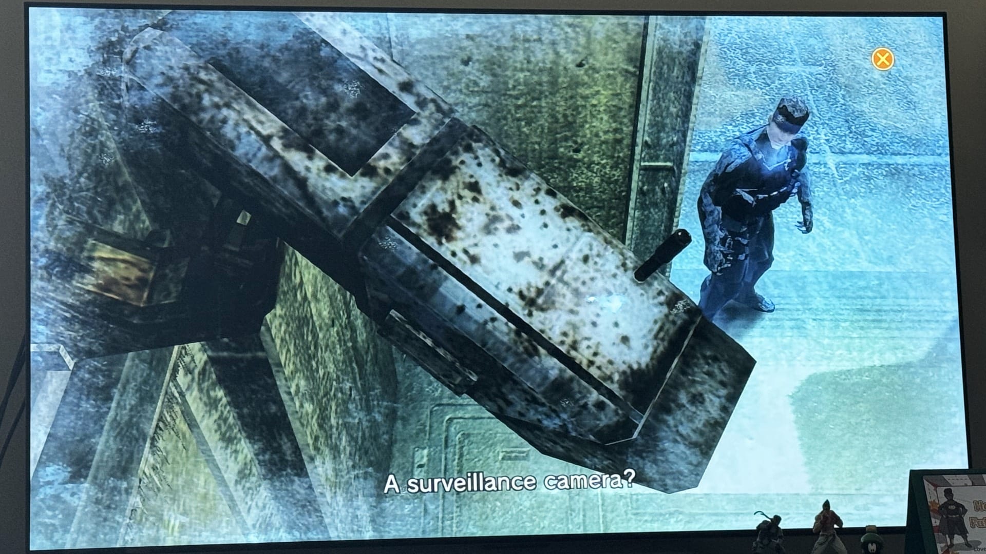 Old Snake looking up at a surveillance camera at Shadow Moses, with the orange X prompt visible in the corner